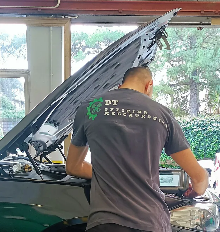 Un meccanico con la maglietta DT Officina Meccatronica controlla il vano motore, con il tablet di diagnosi appoggiato sull'auto